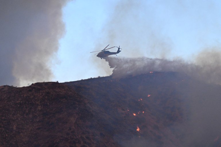 A firefighting helicopter drops water on the Hughes Fire in Castac, a northwest neighborhood of Los Angeles, California on January 22, 2025. On January 22, a new wildfire broke out north of Los Angeles, prompting massive eruptions and evacuation orders for thousands of people in the area. Amazed by the effects of the already huge flames. Terrible flames were consuming hillsides near Caustic Lake, spreading rapidly and engulfing 5,000 acres (2,000 hectares) in just two hours. The fire was being fanned by strong, dry Santa Ana winds that swept across the region, pushing a wide plume of smoke ahead of the flames. About 19,000 people were ordered to evacuate around the lake, which is located 35 miles north of Los Angeles and near the city of Santa Clarita. (Photo by Robin Beck/AFP)