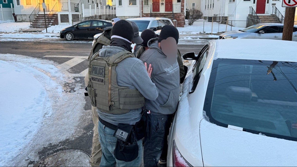 Three ICE officers are standing around a man whose face is blurred when they handcuffs him.