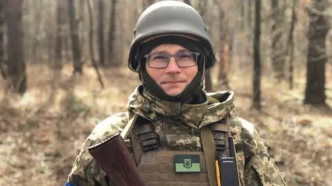 Dimitro Basinkov stands in a forest area dressed in combat gear. He looks directly to the camera. He is wearing a tough armer hat and glasses. You can see that the gun is on top of the gun.