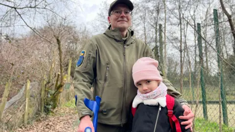 Andrew Marshall/BBC is looking at the camera around a young girl in her left arm. The man is dressed in a khaki green jacket. He is wearing glasses and you can see a small Ukrainian flag on his right arm. The young girl is wearing a pink hat and scarf. You can see the straps of red school bags on his back. You can see the upper part of the blue crusade that the man holds in his right hand.