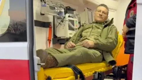 Mermina Basinkov Army is dressed in a yellow stretcher inside an ambulance, dressed in the Army Green Uniform. The man is sitting and wearing glasses.