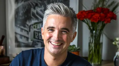 Donal coins have salt and black pepper hair, tied to it and lifted from his forehead. He is wearing a naval T -shirt and a red flower vase is behind where he is sitting