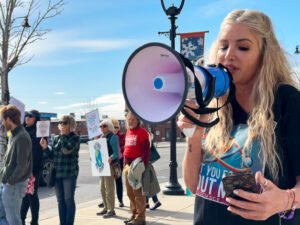 The person wearing a black shirt reads the cell phone and speaks in a megaphone. A group of people stand in the background, which contains signs.