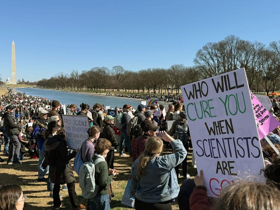 A large crowd gathered in a protest near Washington Memorial, which contains signs of science and education.