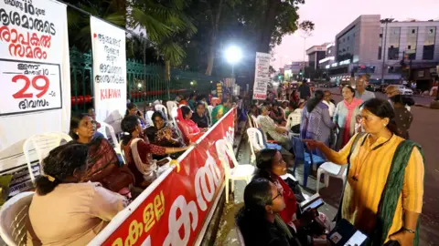 A glimpse of the protest site in front of the Vivek R. Nair/BBC Kerala Secretariat where protesters have camped with play cards and banners on white plastic chairs.