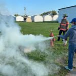 A student uses a fire extinguisher in a grassy field as a firefighter watches