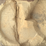 A color close-up photo from the Martian surface shows a circular impression on the flat, hard-looking stone surface, colored brownish-tan, which takes up almost all the image frame. The impression dimensions — its depth compared to its circumference — resembles that of a pie tin. The color of the terrain exposed in the impression is a pale warm tan, much lighter than the surrounding rock. The impression is fractured, in a crack straight down its middle. Another small crack points from the center toward the 4 o’clock position in the circle. To the right of that, the rest of the circle is cleaved off, and the entirety of about one-third of the right side of the circle is missing past a zig-zag line, running from about 1 o’clock to 5 o’clock, leaving an edge that descends below the surface, about twice the depth of the original impression.