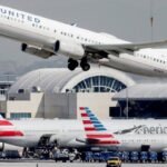 A United Airlines plane is taking off above American Airlines planes parked on the tarmac at Los Angeles International Airport.