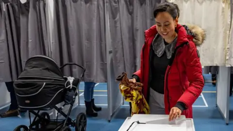 Getty Images A voter dressed in a red coat puts her belt in the tip while her baby waits near the little car